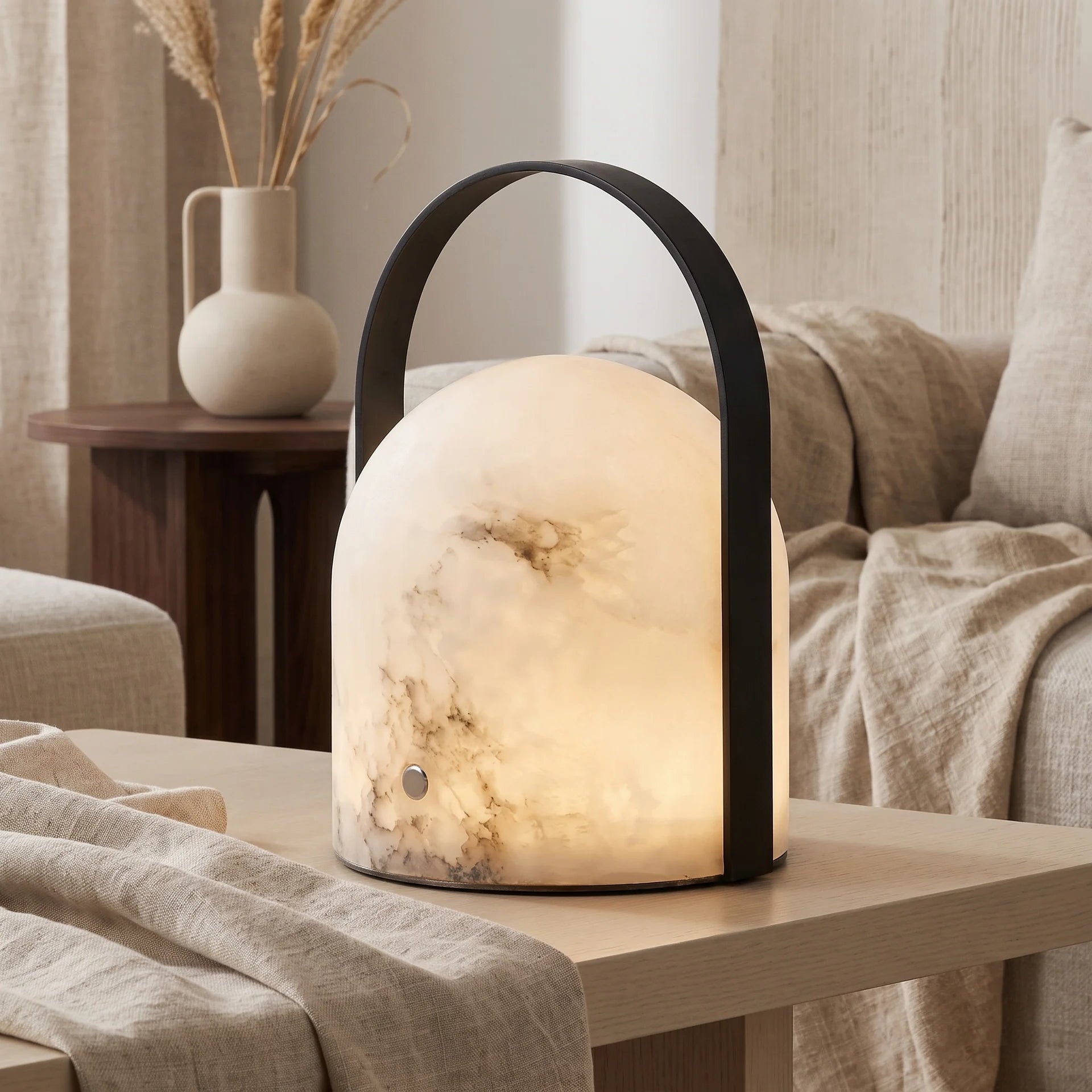 A modern dome-shaped alabaster table lamp with a glowing warm white and light gray marbled body, a black metal handle, and a black base, shown on a wooden table in a cozy living room setting.